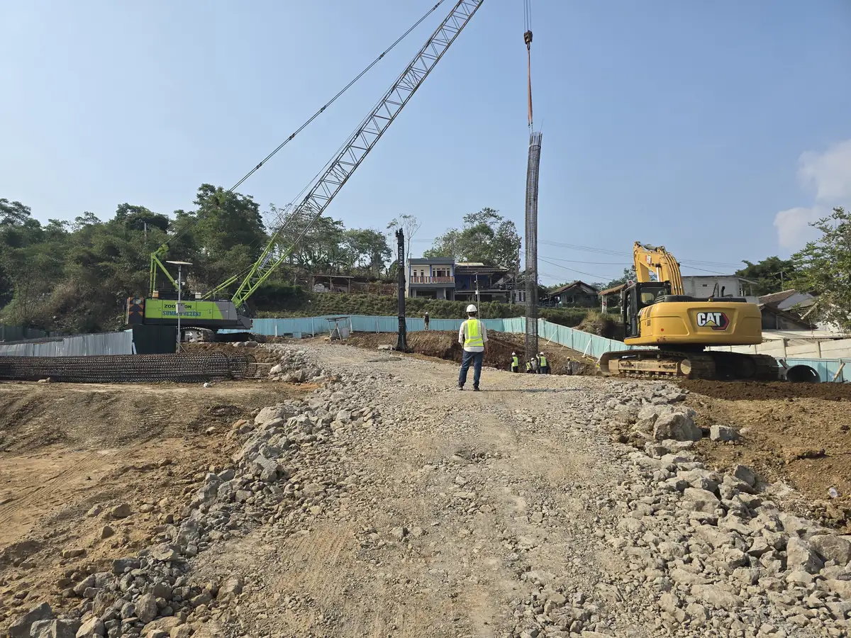 Crane mengangkat keranjang tulangan bored pile di lokasi proyek, dengan jalan kerja dari batu pecah