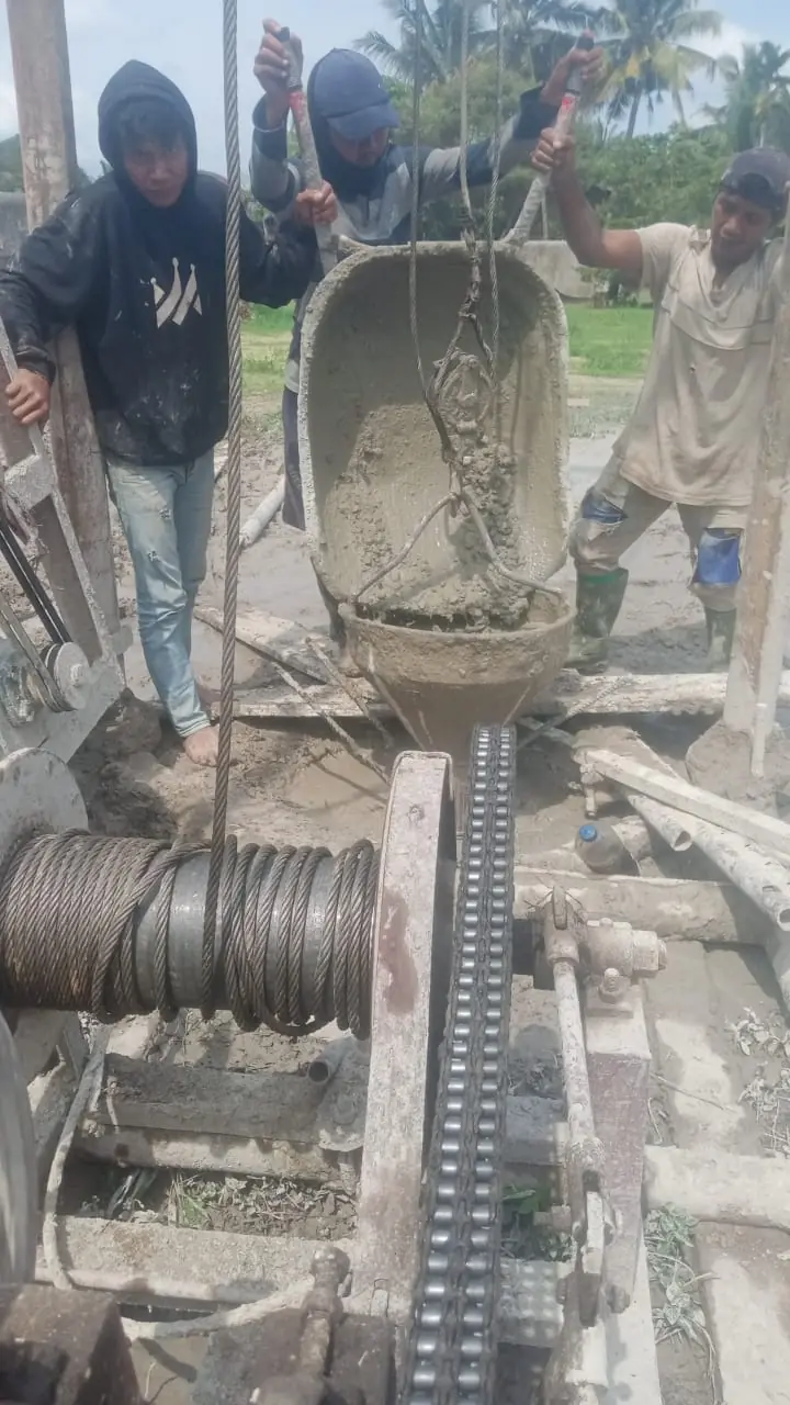 Tiga pekerja mengangkat ember bor berisi tanah basah dari lubang, dengan drum winch di depan