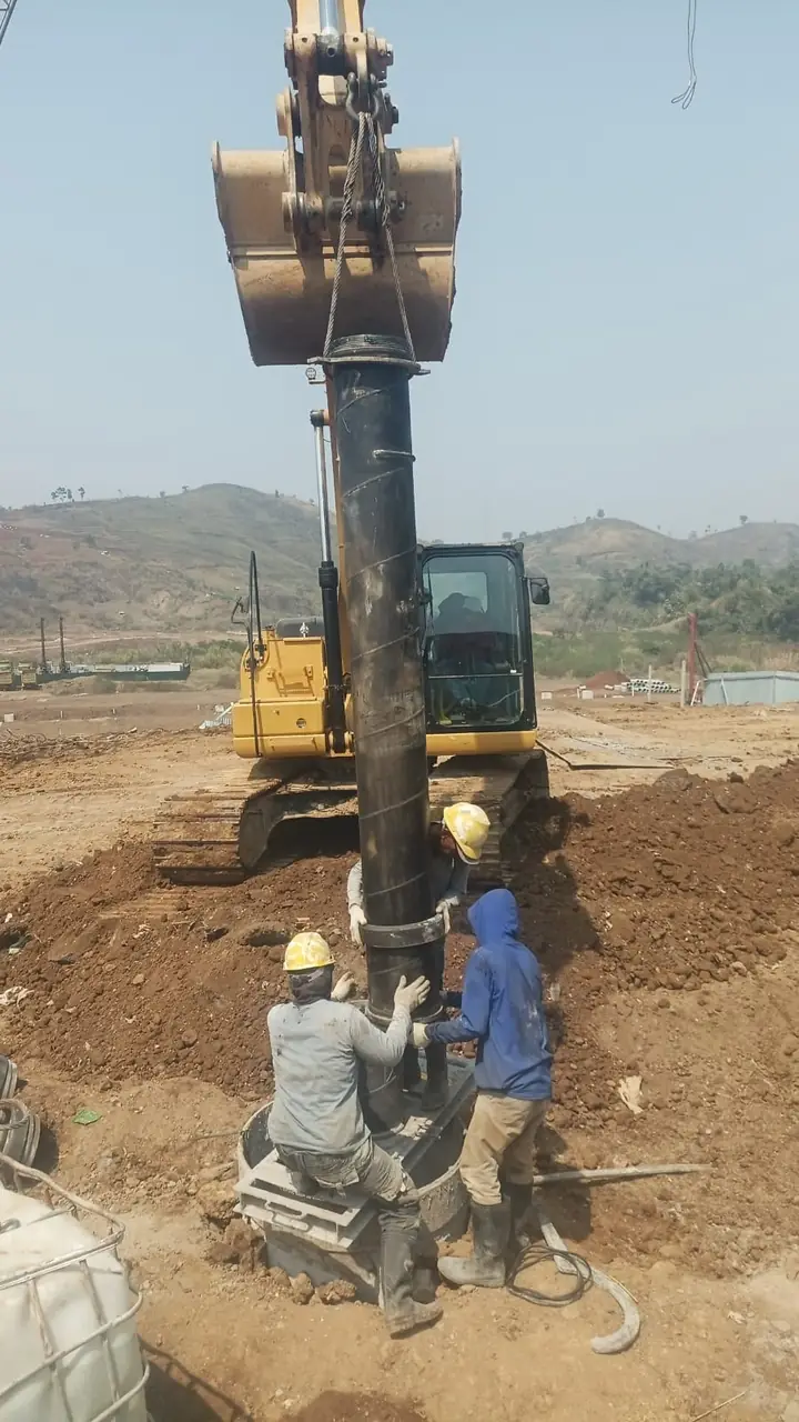 Pipa baja diturunkan ke lubang bor dengan bantuan excavator, dipandu dua pekerja