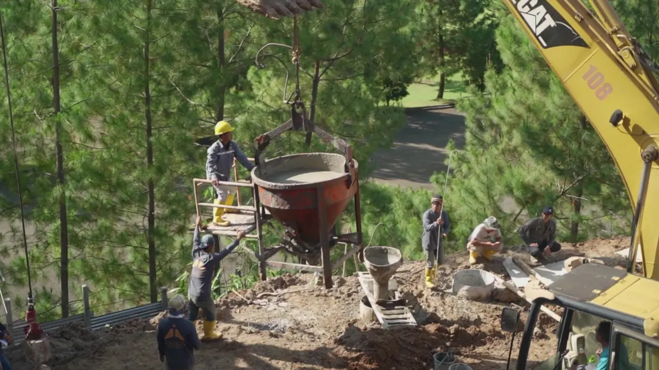 Workers discharging concrete from a crane-suspended bucket into a pile position among pine trees