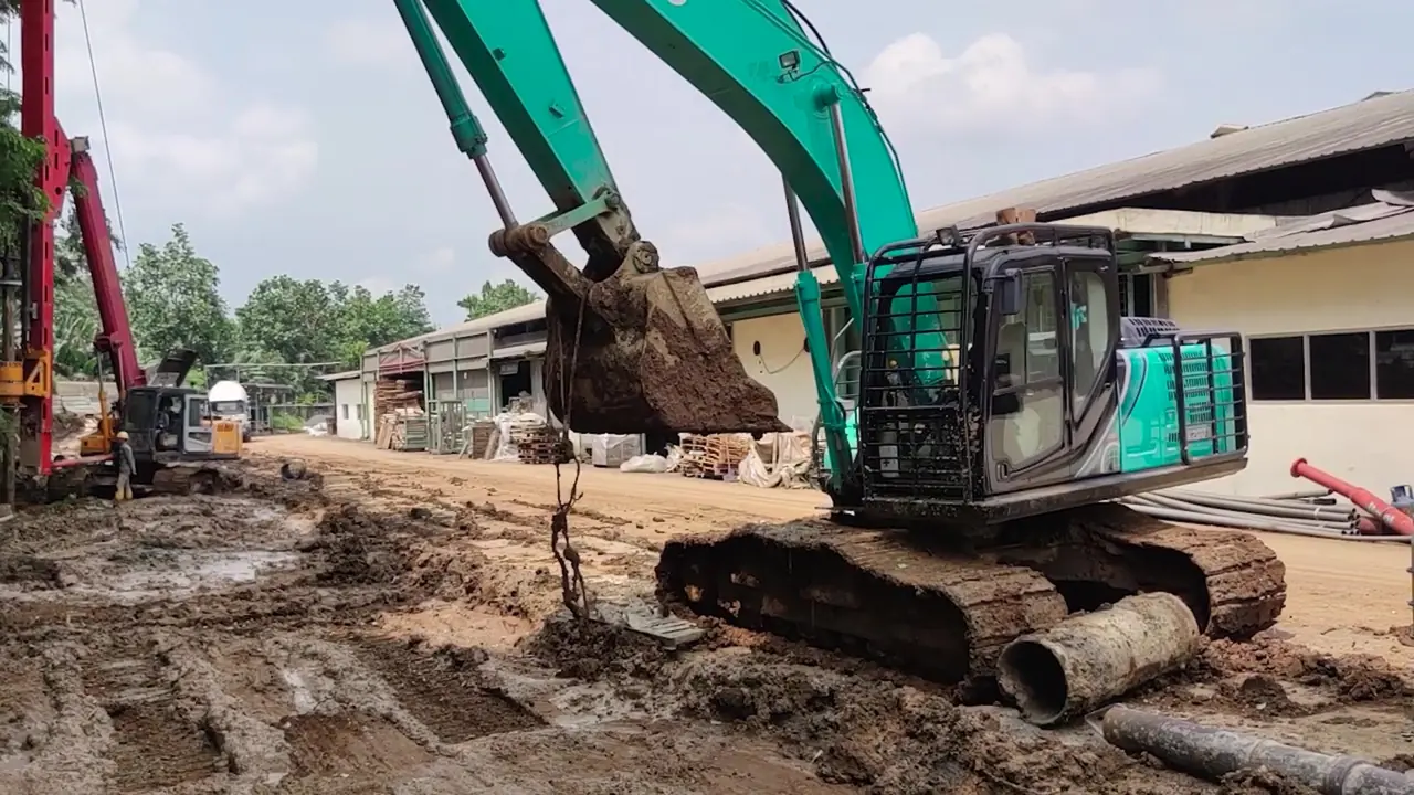 Excavator mengeruk di jalur kerja sempit di antara bangunan pabrik Indah Kiat yang tetap beroperasi, dengan rig full casing di kejauhan