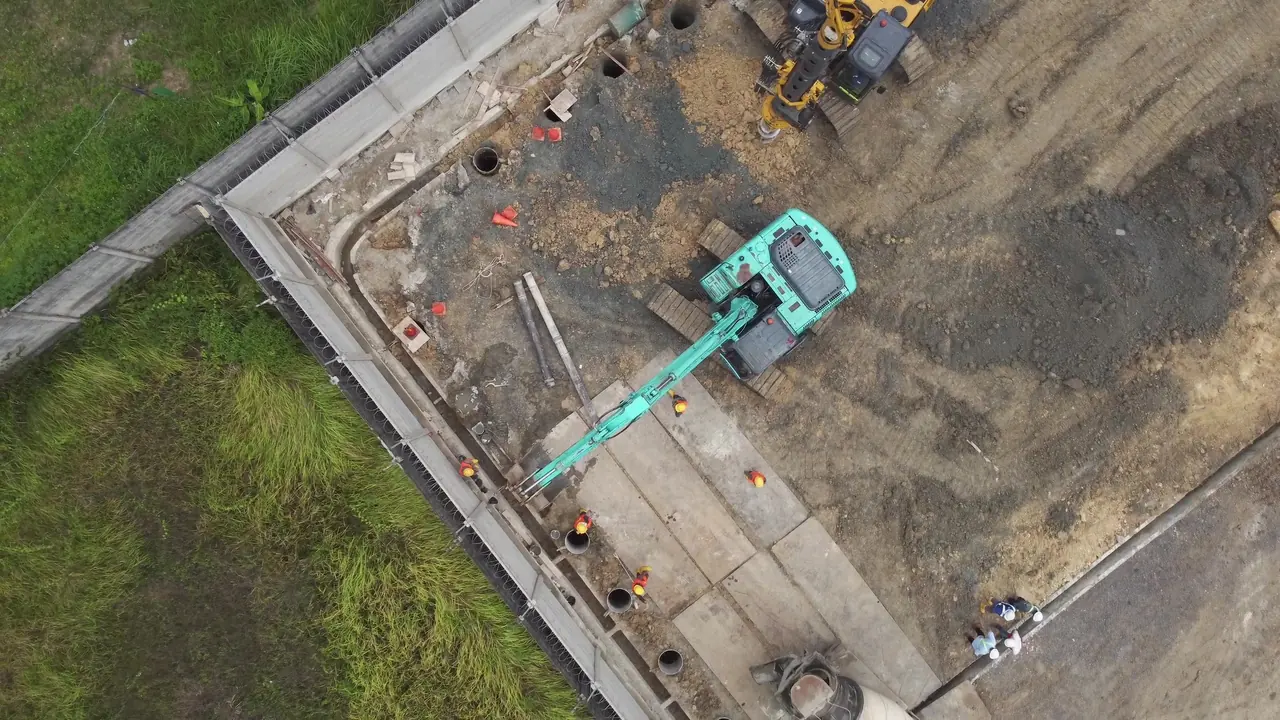 Foto udara tegak lurus barisan lubang bor dan pipa casing di sepanjang pagar batas lahan, dengan excavator dan truk mixer di jalur kerja