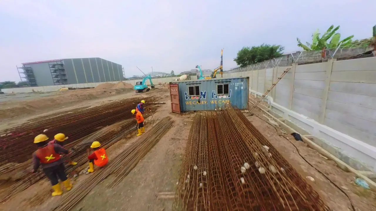 Pekerja merakit keranjang tulangan yang tersusun berjajar di area fabrikasi, dengan rig bor dan truk mixer di latar belakang