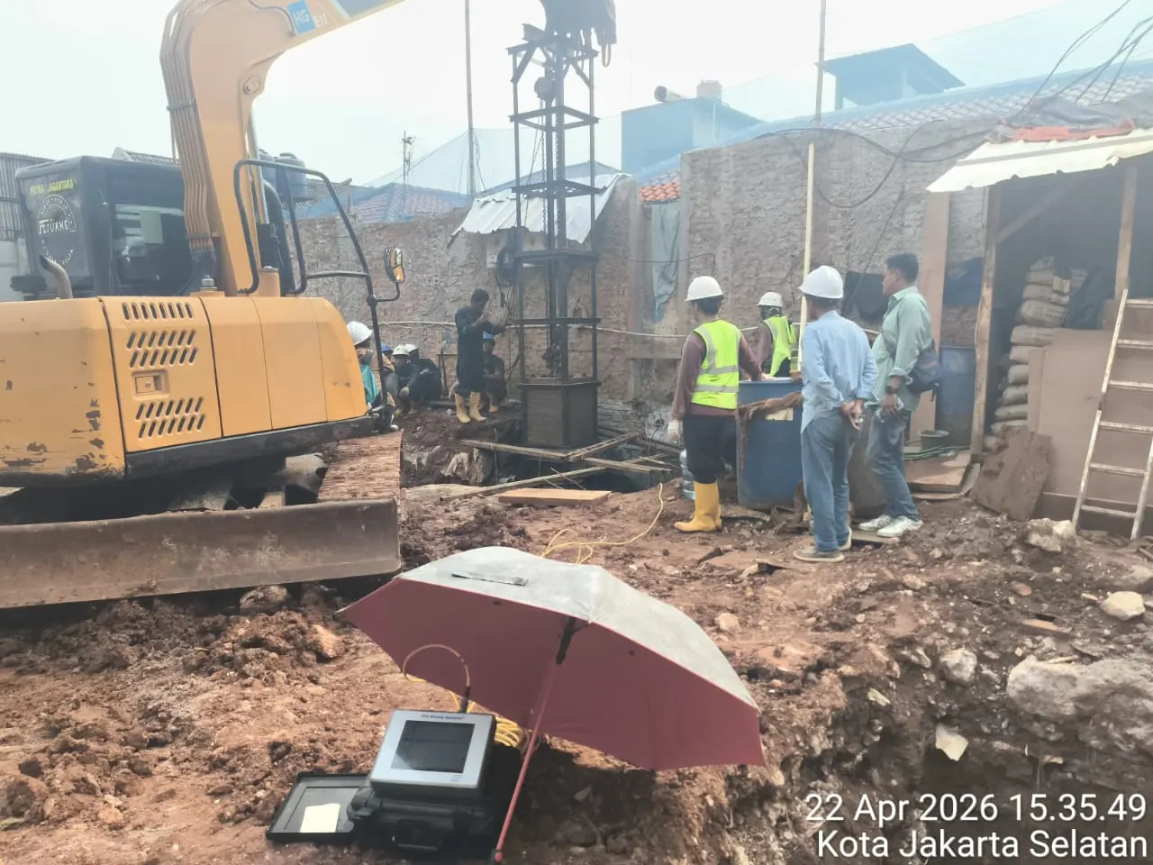 Perangkat Pile Driving Analyzer berteduh di bawah payung di tengah lahan, dengan excavator dan tim lapangan di belakangnya